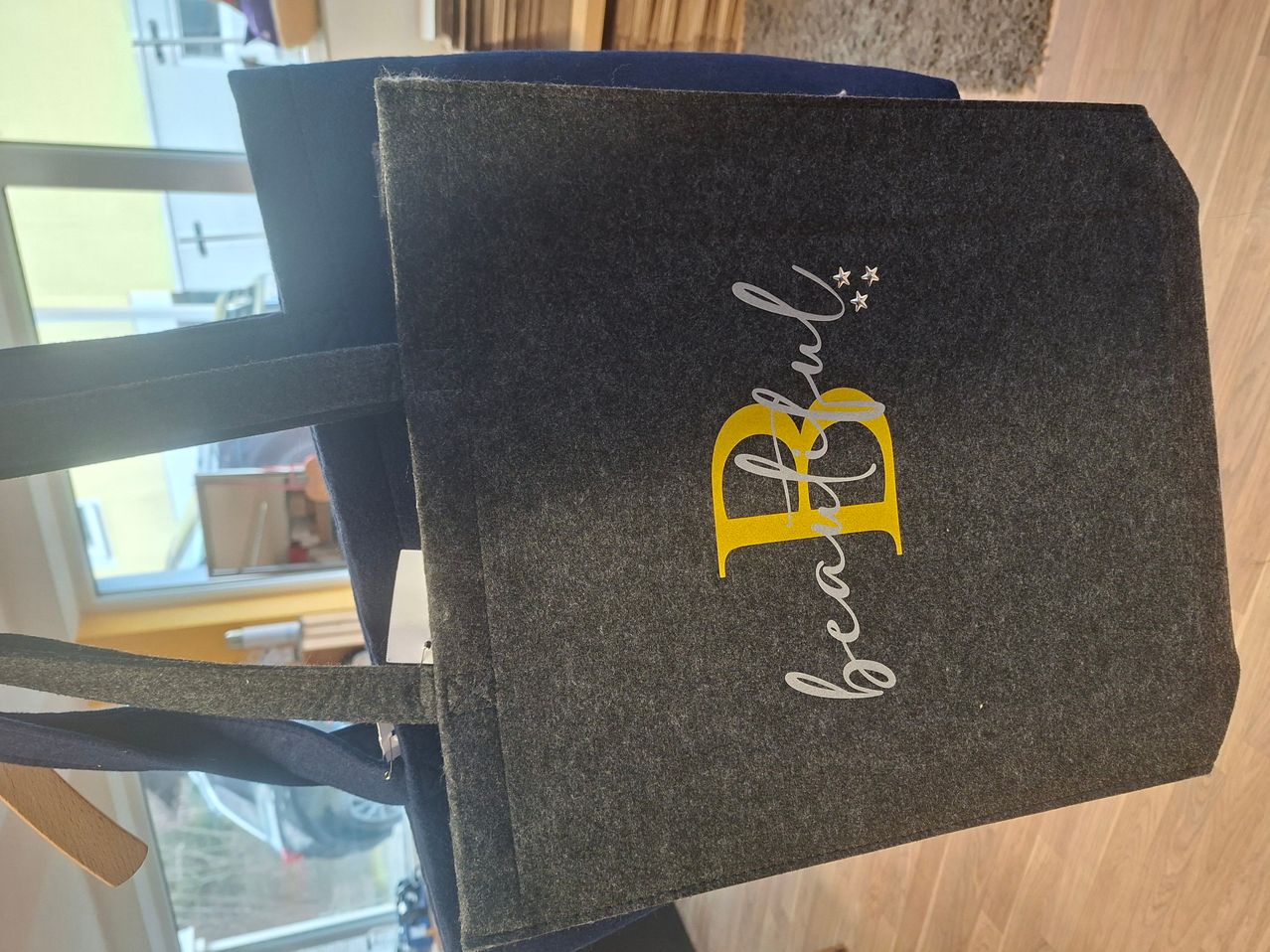 Eine graue Tragetasche mit dem Wort 'Geburtstag' und einem gelben Herzlogo mit Sternen darüber. Sie liegt auf einem blauen Stuhl.