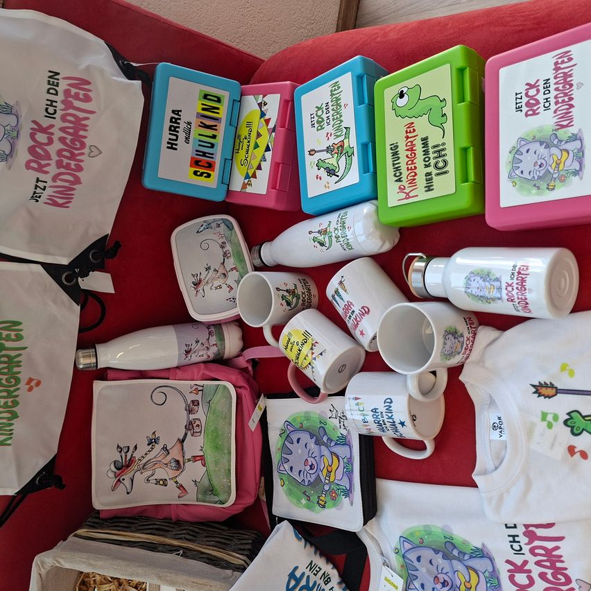 A collection of colorful items including lunch boxes, mugs, and bags with the word 'kindergarten' on them. The items are arranged on a red surface.