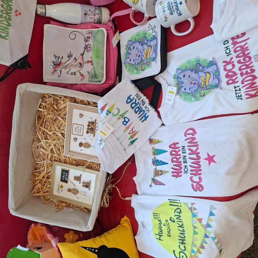 A collection of items on a red surface, including a box with straw, a pink bag, mugs, and shirts with cartoon cat designs.