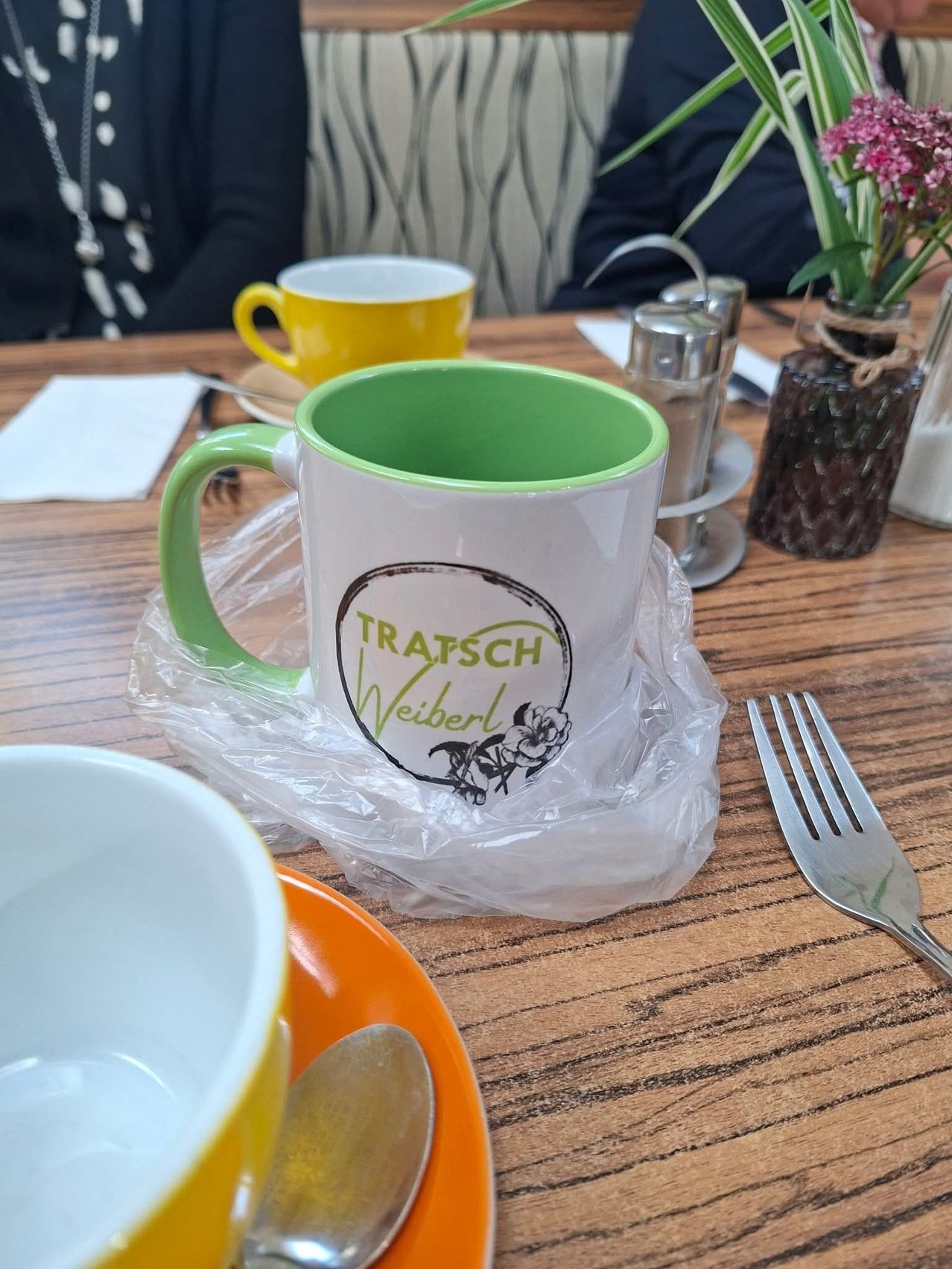 A wooden table with a green and white mug labeled 'TRATSCH Weibert', a yellow mug, a vase with pink flowers, a fork, and a spoon.