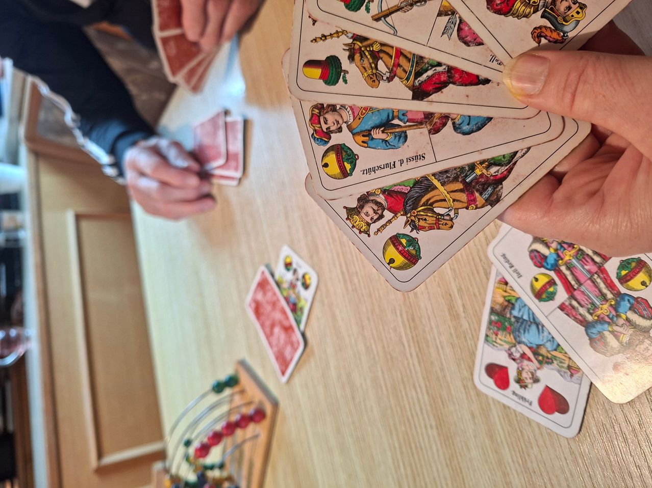 Eine Person hält ein Kartenspiel mit Illustrationen historischer Figuren, während eine andere Person eine Karte in der Hand hält. Die Karten liegen auf einem Holztisch.