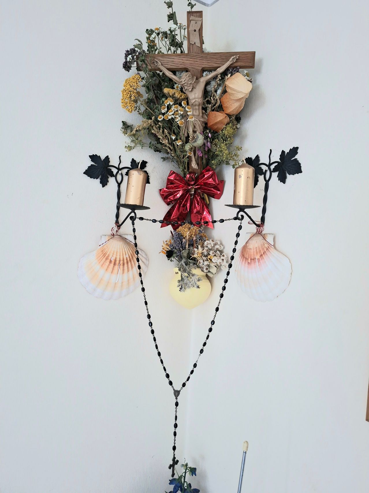 Eine religiöse Wanddekoration mit einem Kruzifix, Kerzen, Blumen und Muschelschalen auf weißem Hintergrund.