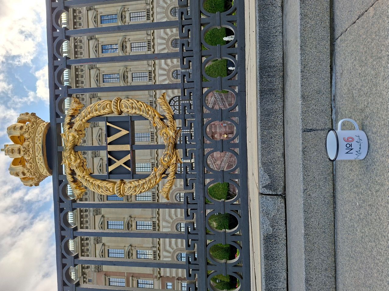 Ein goldener Lorbeerkranz mit den Buchstaben 'XX' in der Mitte ist an einem schwarzen Metallzaun angebracht. Hinter dem Zaun ist ein Gebäude mit vielen Fenstern und einer Ziegelmauer zu sehen.