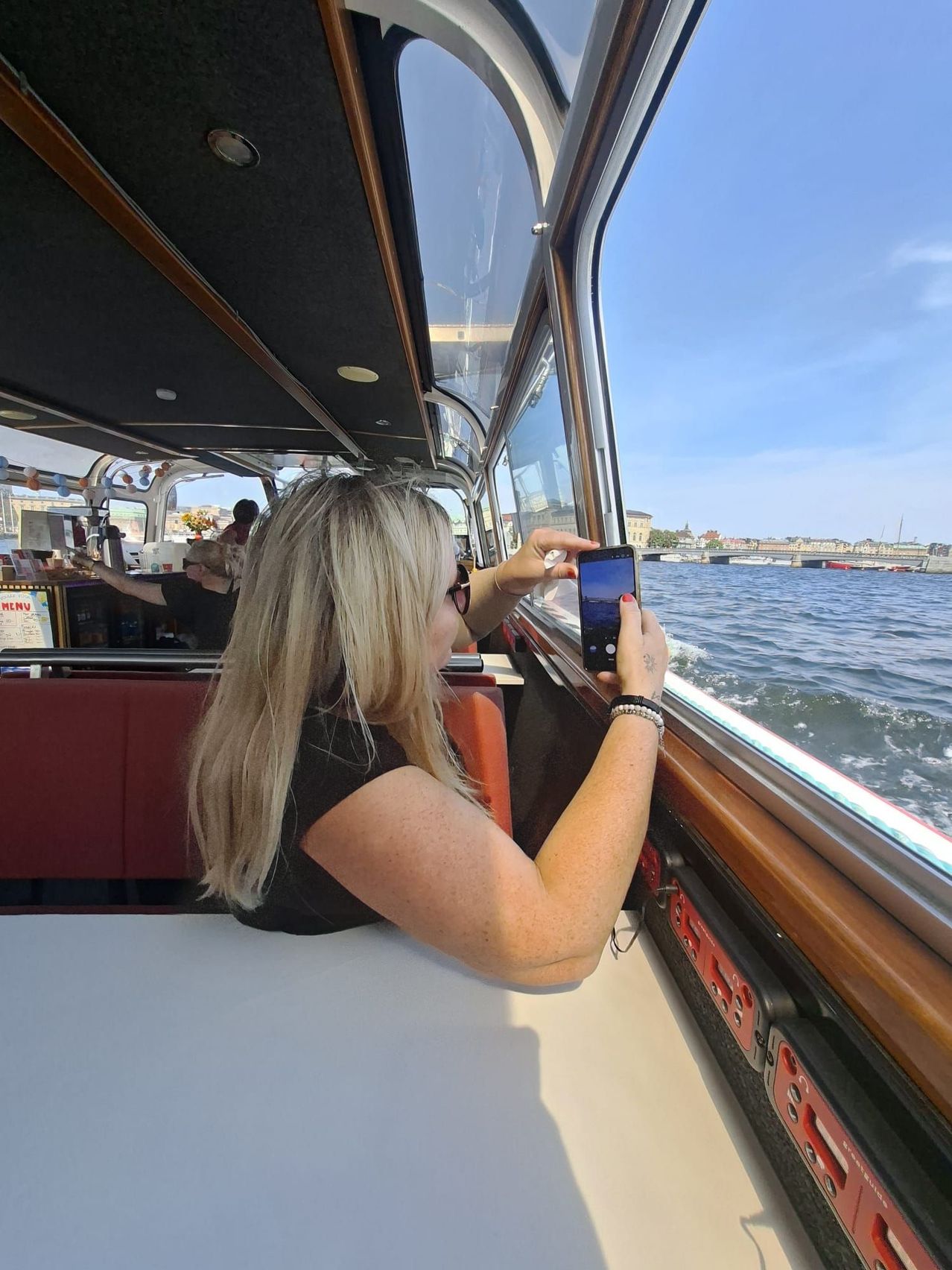 Eine Frau auf einem Boot macht ein Foto vom Wasser mit ihrem Handy. Sie trägt eine Sonnenbrille und ein schwarzes Shirt.