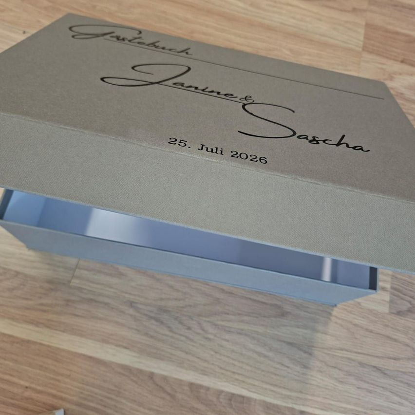 A gray box with the names Janine and Sascha and the date July 25, 2026, placed on a wooden surface.