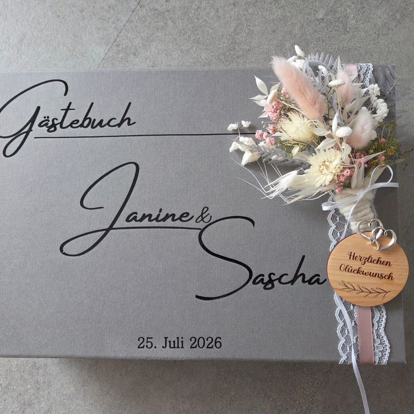 A gray guest book with the names Janine and Sascha written in cursive, adorned with a pink and white floral arrangement and a wooden heart tag that reads 'Herzlichen Gluckwunsch'. The date 25th July 2026 is also visible.