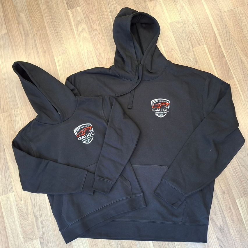 Zwei schwarze Kapuzenpullover mit weißen Logos liegen auf einem Holzboden. Der größere Pullover hat ein rotes Logo, während der kleinere ein weißes Logo hat.