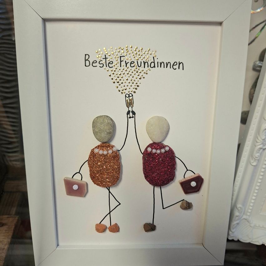 A framed artwork featuring two women holding hands, with a golden sparkle above them and the text 'Beste Freundinnen' written in German. The women are made from stones and glitter.