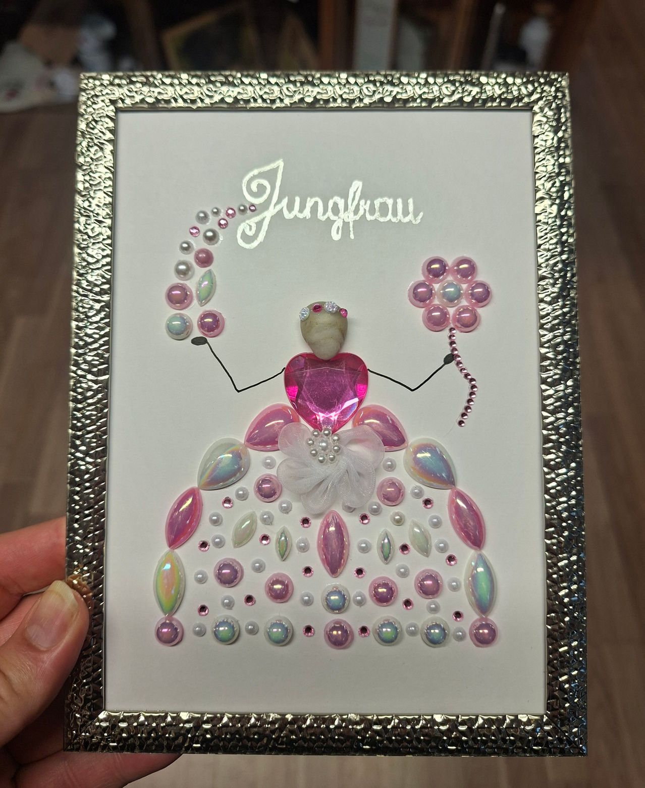 A framed art piece featuring a girl with a pink dress adorned with various pink and white beads, set against a white background. The word 'Jungfrau' is written in white at the top.