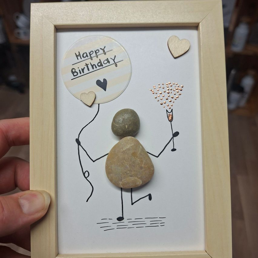A framed art piece featuring a stick figure holding a balloon with 'Happy Birthday' written on it, a rock body, and a champagne flute. The artwork is surrounded by hearts and is held by a person.