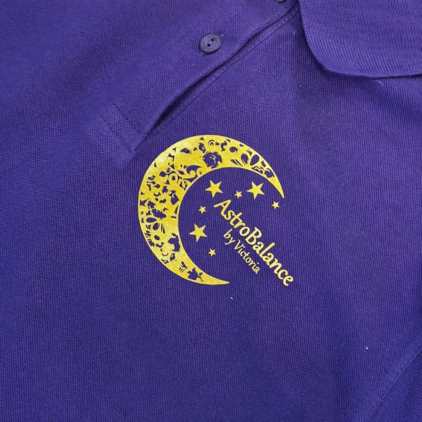 A close-up of a purple polo shirt featuring a yellow logo with a crescent moon, stars, and the words 'AstroBalance by Victoria'.