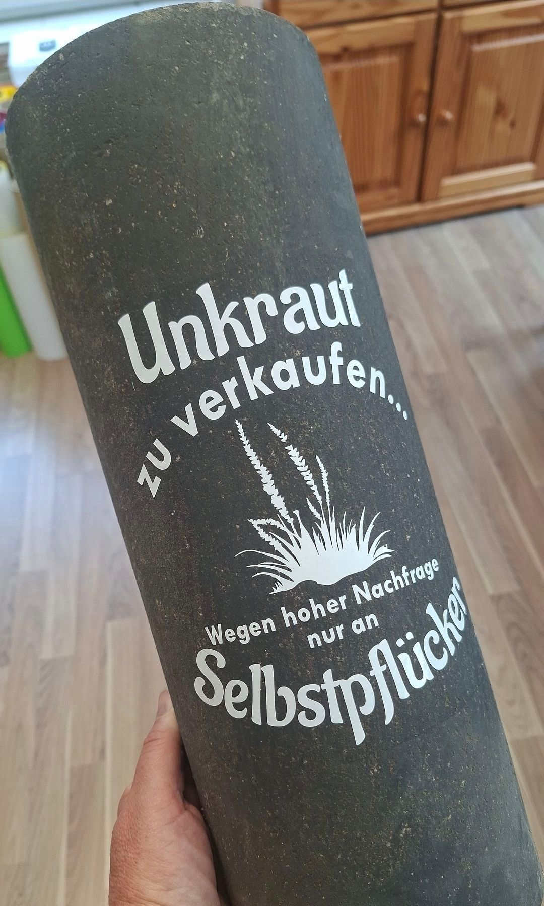 Ein schwarzer zylindrischer Gegenstand mit weißem Text. Es steht 'Unkraut zuverkaufen' mit einer Pflanzenillustration. Darunter steht 'Wegen hoher Nachfrage nur an Selbstpflücker'. Es scheint ein Behälter oder eine Verpackung für Unkraut oder Kräuter zu sein.