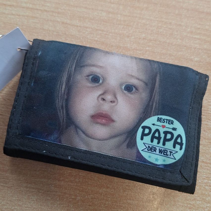 Ein Portemonnaie mit einem Foto eines kleinen Mädchens, beschriftet mit 'Bester Papa der Welt', auf einer Holzoberfläche platziert.