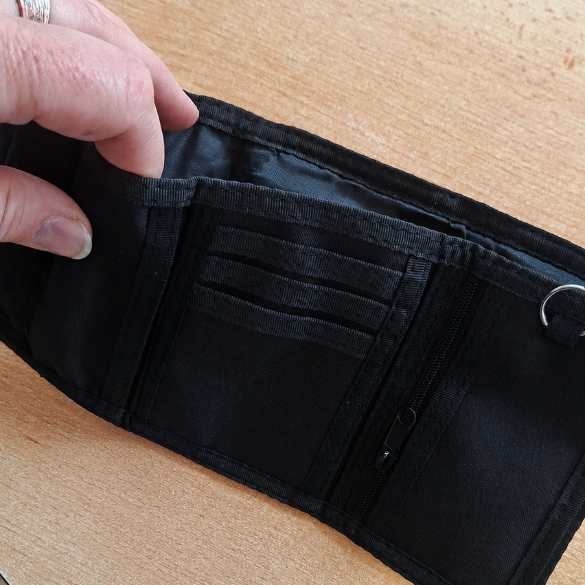 Eine Hand öffnet ein schwarzes Portemonnaie mit Reißverschlüssen und Taschen auf einem Holztisch.
