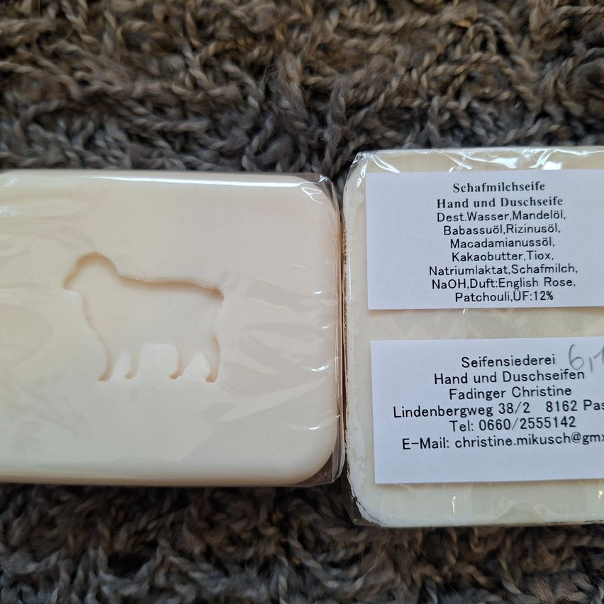 Two handmade soap bars with animal shapes, one with a horse and the other with a sheep, placed on a textured surface. Labels indicate they are hand and shower soaps with various natural ingredients.
