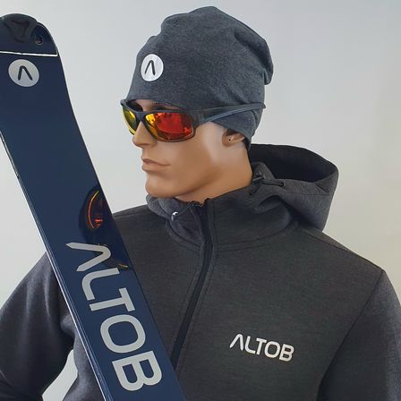Ein Mannequin trägt eine graue Mütze, eine Sonnenbrille und einen schwarzen Kapuzenpullover mit weißen Buchstaben 'ALTO'. Es zeigt einen blauen Snowboard mit dem Wort 'ALTO'.