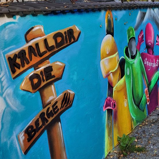 Ein farbenfrohes Wandbild mit einem Schild, auf dem 'Kralldir Die Berge' steht, an einer blauen Wand mit einer kleinen Pflanze am Boden.