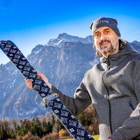 Ein Mann mit einem lächelnden Gesicht hält ein Snowboard in beiden Händen. Er trägt eine graue Kapuzenjacke. Hinter ihm befindet sich eine Bergkette. Der Himmel ist klar.