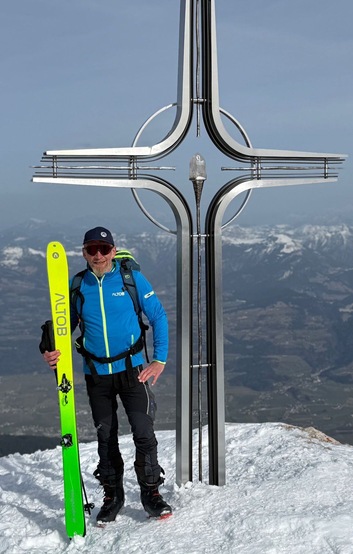 Ein Mann in einem blauen Jackett und schwarzen Hosen steht auf einem verschneiten Gipfel und hält einen gelben Ski mit dem Wort Altob. Er steht neben einem großen metallischen Kreuz.