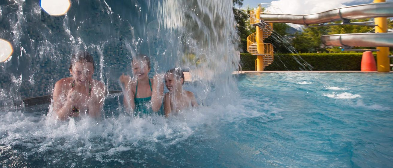 Drei Kinder stehen unter einem Wasserfall im Pool und lächeln. Dahinter befindet sich ein gelber Spiralrutsche.