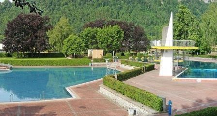Ein Freibad mit Wasserrutsche, umgeben von gepflegten Hecken, Bäumen und Bergen im Hintergrund.