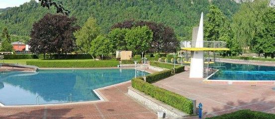Ein Freibad mit Wasserrutsche, umgeben von gepflegten Hecken, Bäumen und Bergen im Hintergrund.