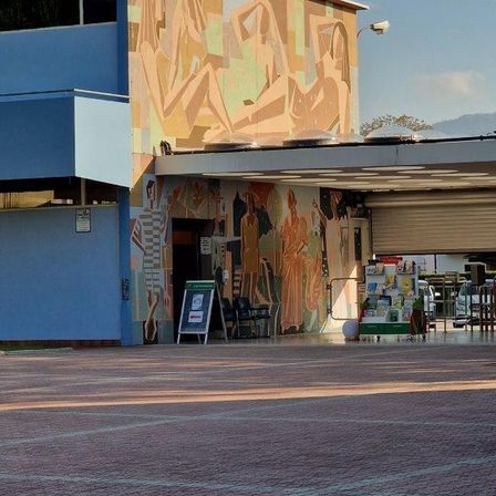 Ein blaues Gebäude mit einem großen Wandbild an der Wand und einem Schild davor. Das Wandbild zeigt verschiedene Figuren und ein geparktes Auto.