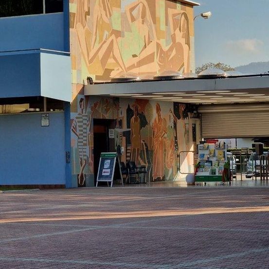 Ein blaues Gebäude mit einem großen Wandbild an der Wand und einem Schild davor. Das Wandbild zeigt verschiedene Figuren und ein geparktes Auto.