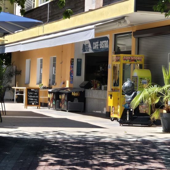 Ein gelbes Geschäft mit einem weißen Vordach trägt die Aufschrift 'CAFE BISTRO'. Ein schwarzes Tafelmenü ist draußen aufgestellt. Ein Spielautomat steht vor dem Gebäude. Ein Topfpalme steht rechts.