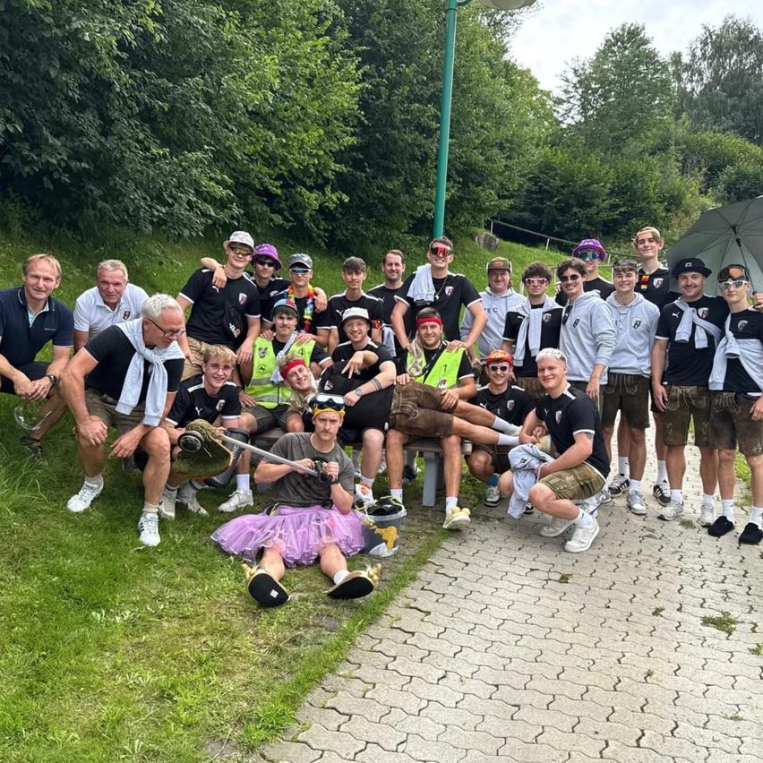 Eine Gruppe von Menschen in Sportkleidung posiert für ein Foto auf einem gepflasterten Weg, einige sitzend und andere stehend, in einem Außenbereich mit Bäumen und einer Straßenlaterne.