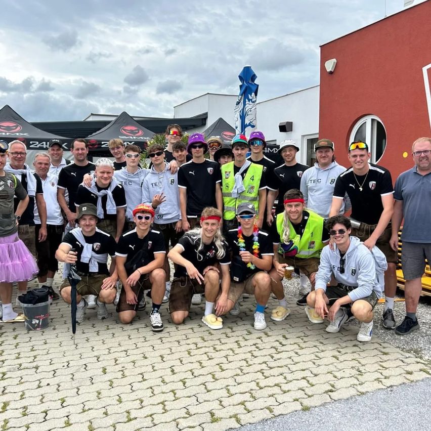 Eine Gruppe von Menschen in verschiedenen farbenfrohen und thematischen Kostümen posiert für ein Foto auf einer Kopfsteinpflasterstraße. Zelte sind im Hintergrund zu sehen.