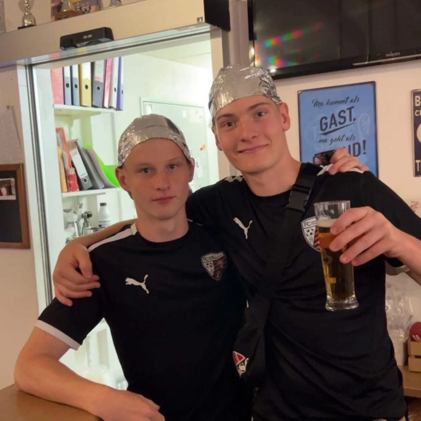 Zwei Männer mit Aluminiumfolien-Hüten und schwarzen Trikots mit weißen Logos posieren mit einem Glas Bier. Dahinter steht eine Glastür mit Regalen, einem Fernseher und einem blauen Plakat.