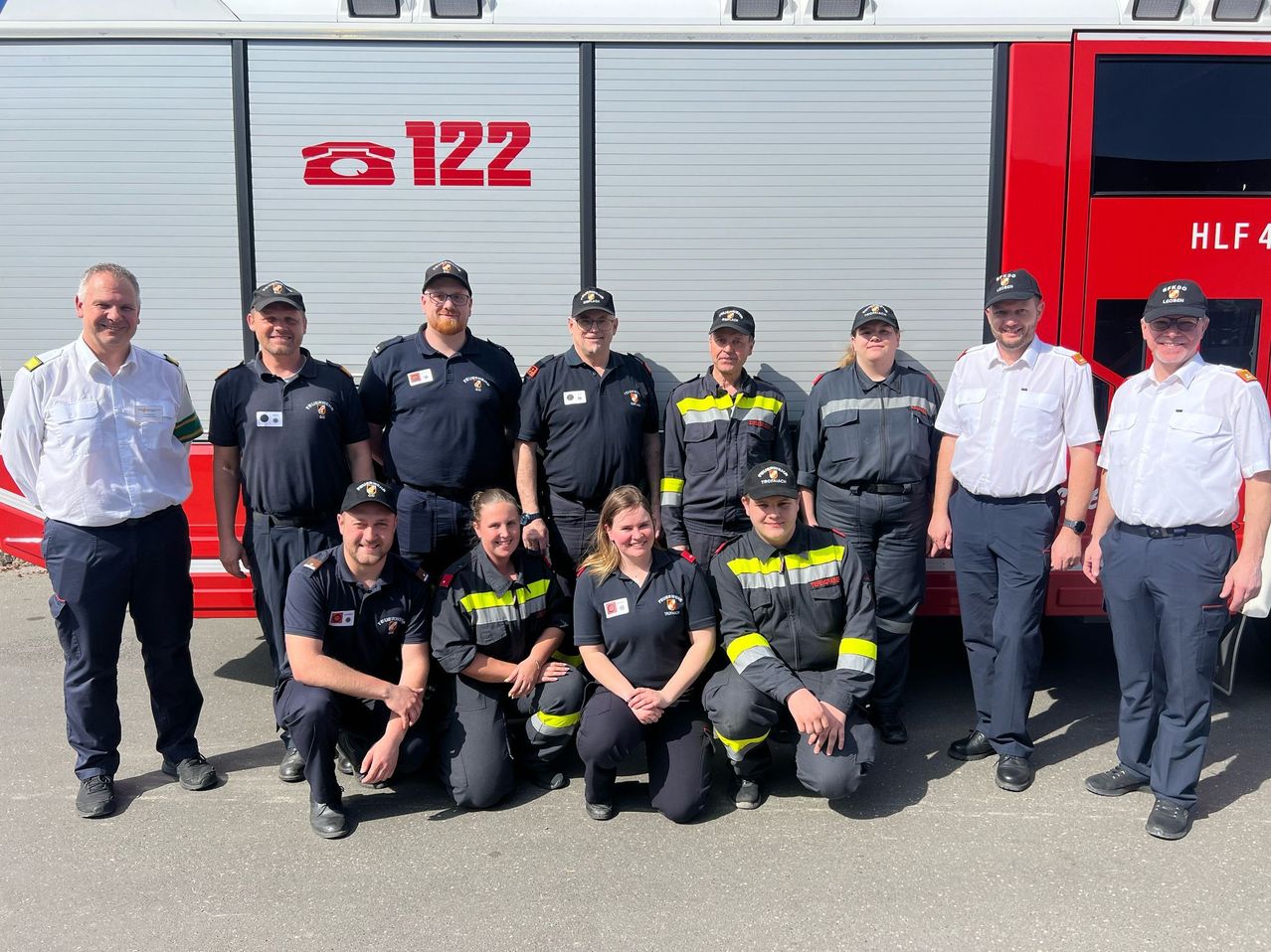 Eine Gruppe Feuerwehrleute, sowohl Männer als auch Frauen, steht und kniet vor einem roten Feuerwehrauto. Sie tragen Uniformen und Mützen, und einige von ihnen lächeln.