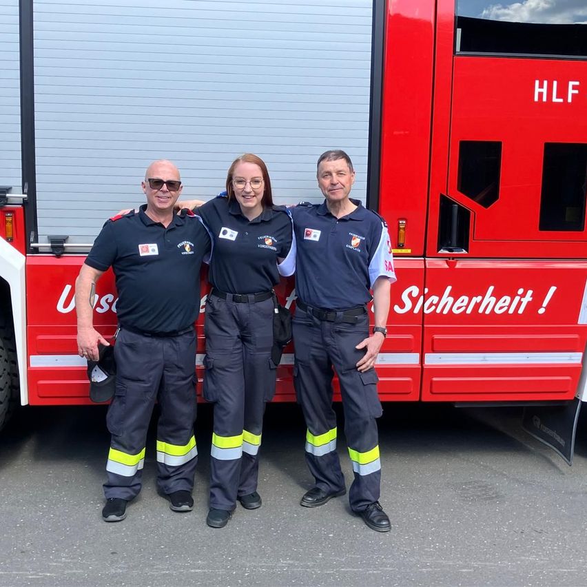 Drei Personen in Uniform stehen vor einem roten Feuerwehrfahrzeug mit der Aufschrift 'HLF' und 'Sicherheit!'.