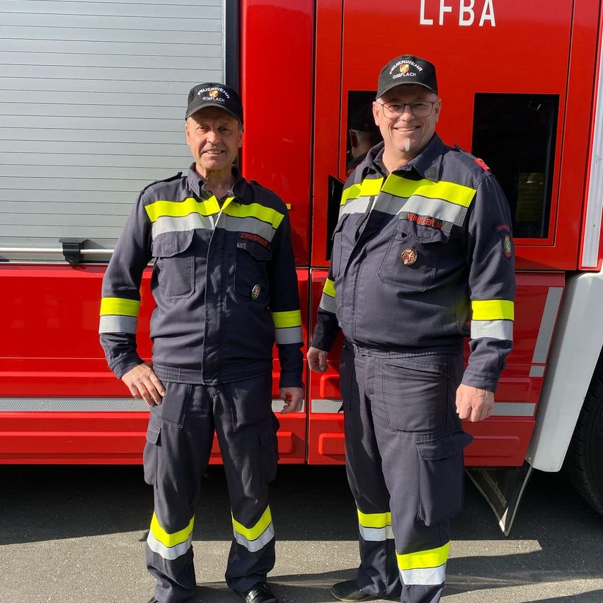 Zwei Feuerwehrleute in Uniform stehen vor einem roten Feuerwehrwagen.