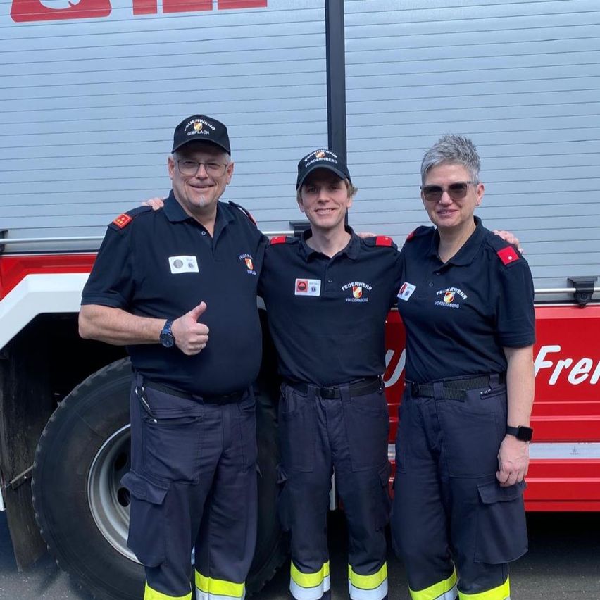 Drei Feuerwehrleute stehen vor einem Feuerwehrauto. Der Mann auf der linken Seite gibt einen Daumen hoch. Der Mann in der Mitte trägt einen Hut. Die Frau auf der rechten Seite trägt eine Brille.