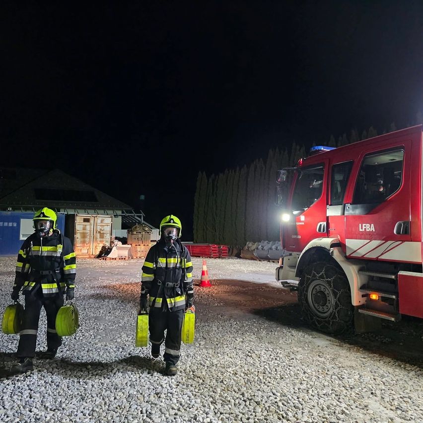 Zwei Feuerwehrleute stehen vor einem roten Feuerwehrauto mit den Buchstaben LFBA, beide tragen gelbe Helme und tragen gelbe Ausrüstung.