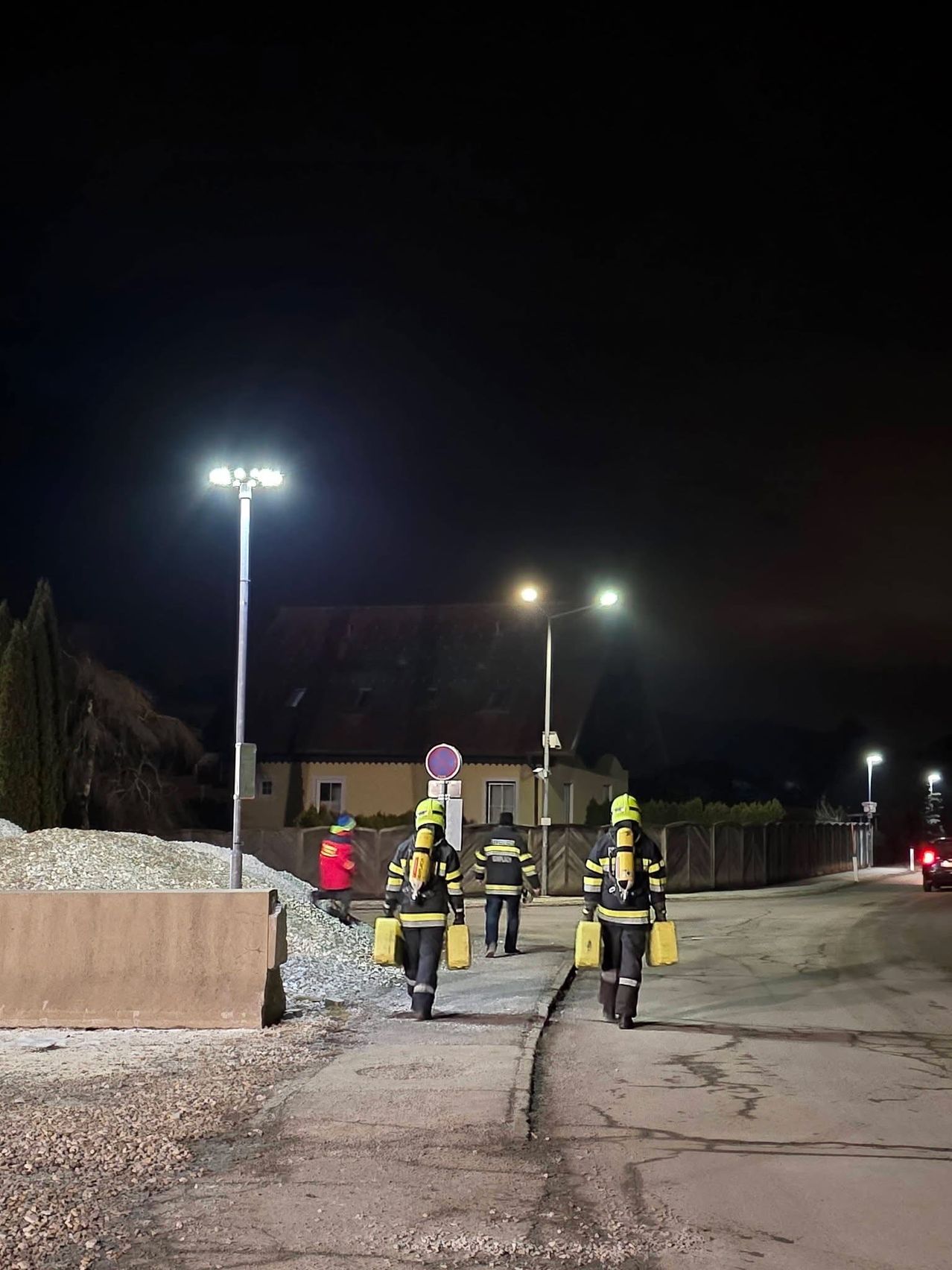 Drei Feuerwehrleute mit gelben Helmen gehen bei Nacht mit gelber Ausrüstung die Straße entlang. Eine Person in einer roten Jacke steht neben einem Haufen Kies. Straßenlaternen und ein Gebäude sind sichtbar.