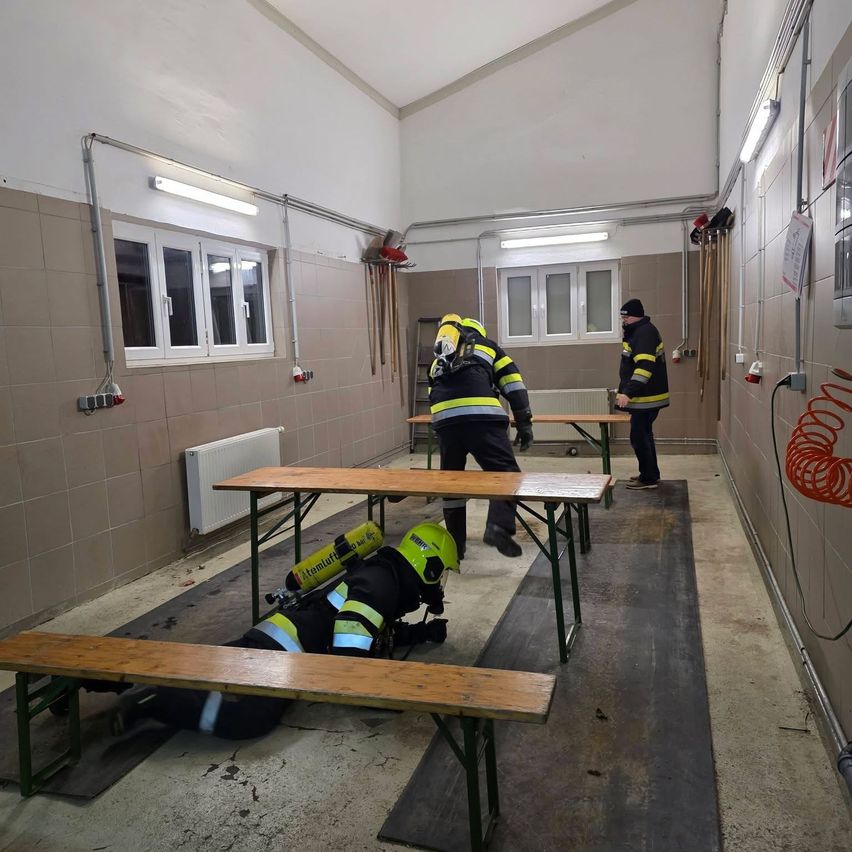 Drei Feuerwehrleute üben in einem Raum mit Tischen, Bänken und einem Heizkörper. Zwei stehen und einer liegt auf dem Boden. An der Wand sind Werkzeuge montiert.