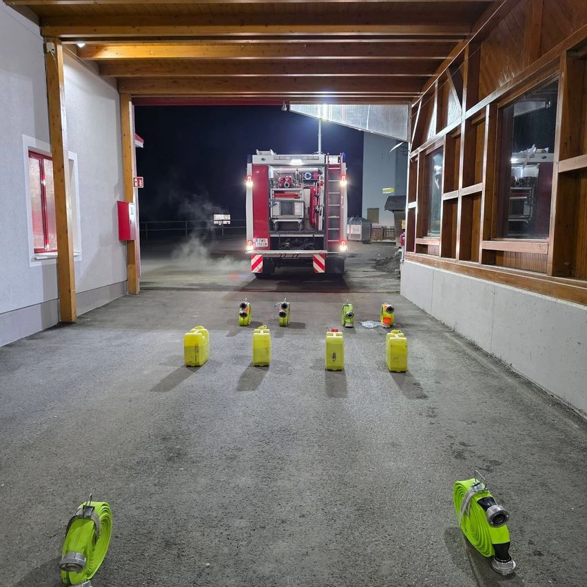 Ein Feuerwehrauto steht nachts in einer Garage. Es ist beleuchtet und Rauch kommt heraus. Mehrere gelbe Objekte sind in einer Reihe auf dem Boden angeordnet.