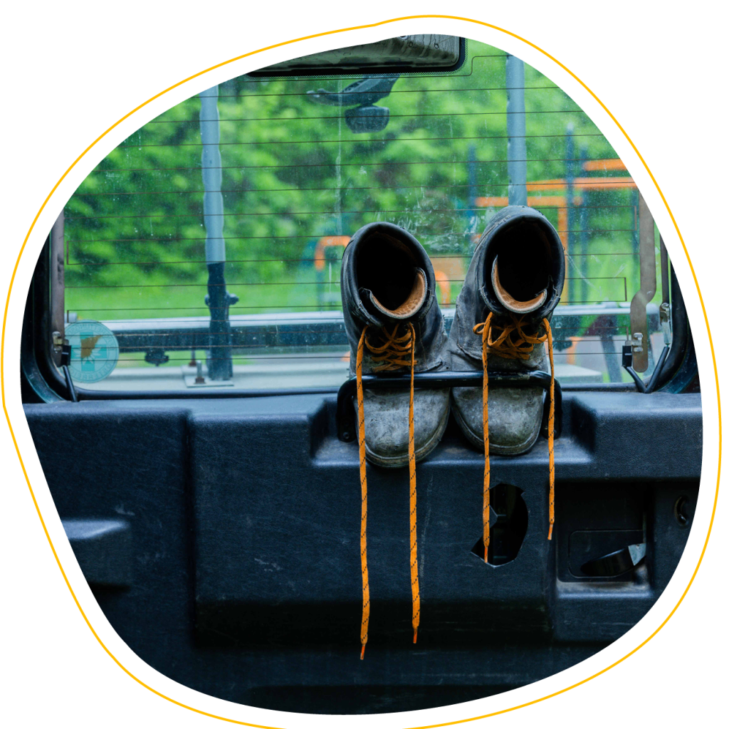 A pair of worn boots hang from the back seat of a vehicle, with a view of a park through the window.