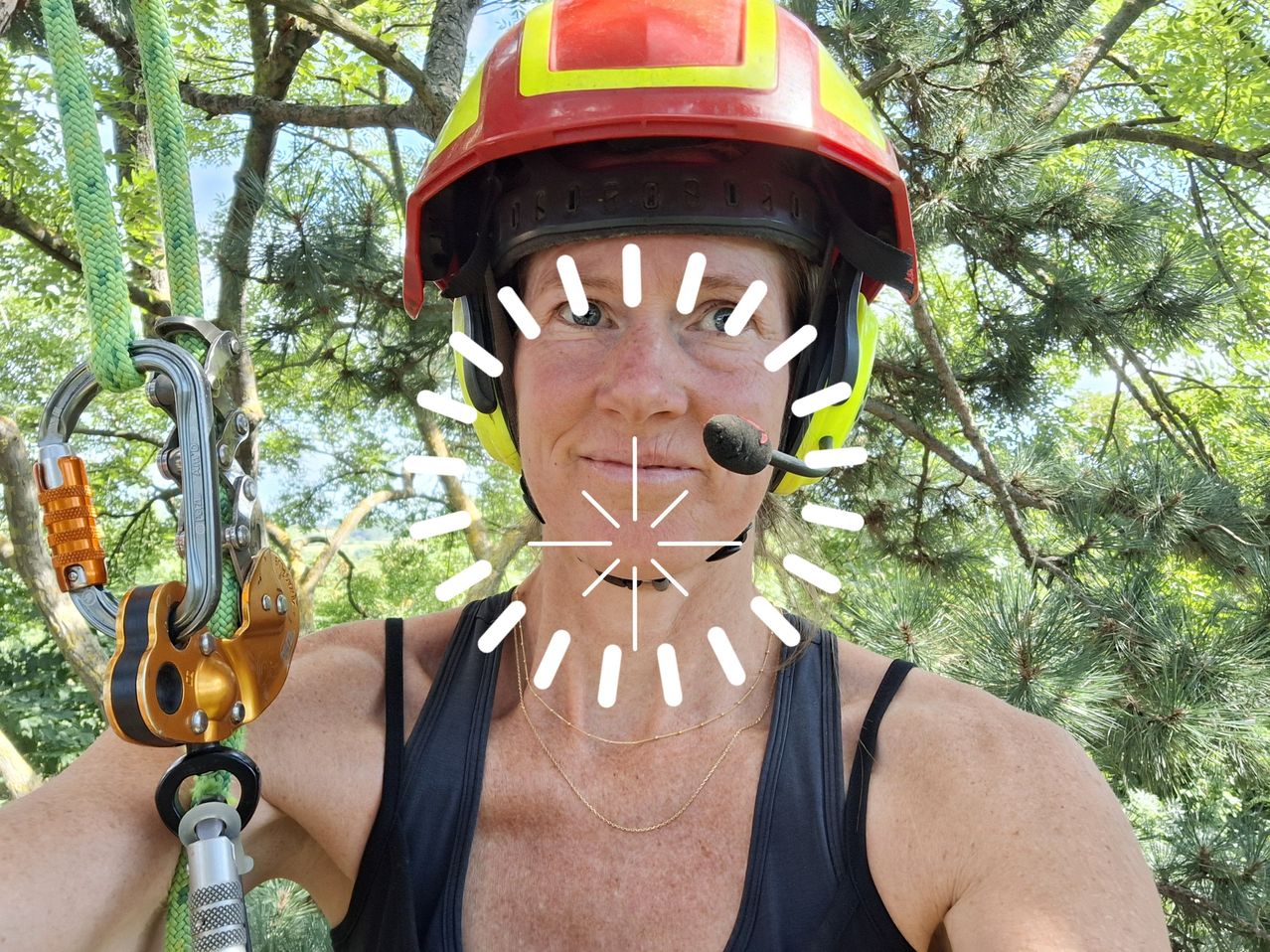 A woman in a red and yellow helmet with a microphone in her mouth is standing in a forest.