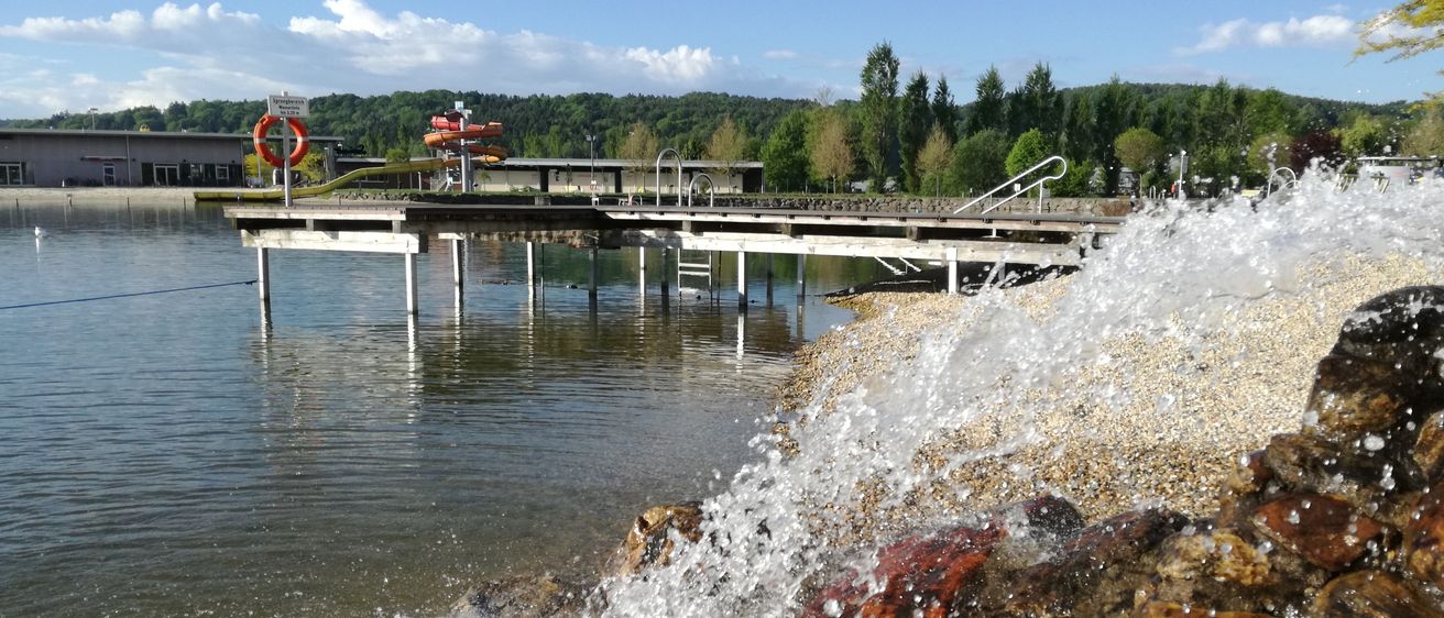 Eine Anlegestelle mit einer Rutsche ragt in einen See, umgeben von Bäumen und Bergen unter einem bewölkten Himmel. Wasser spritzt um Steine.