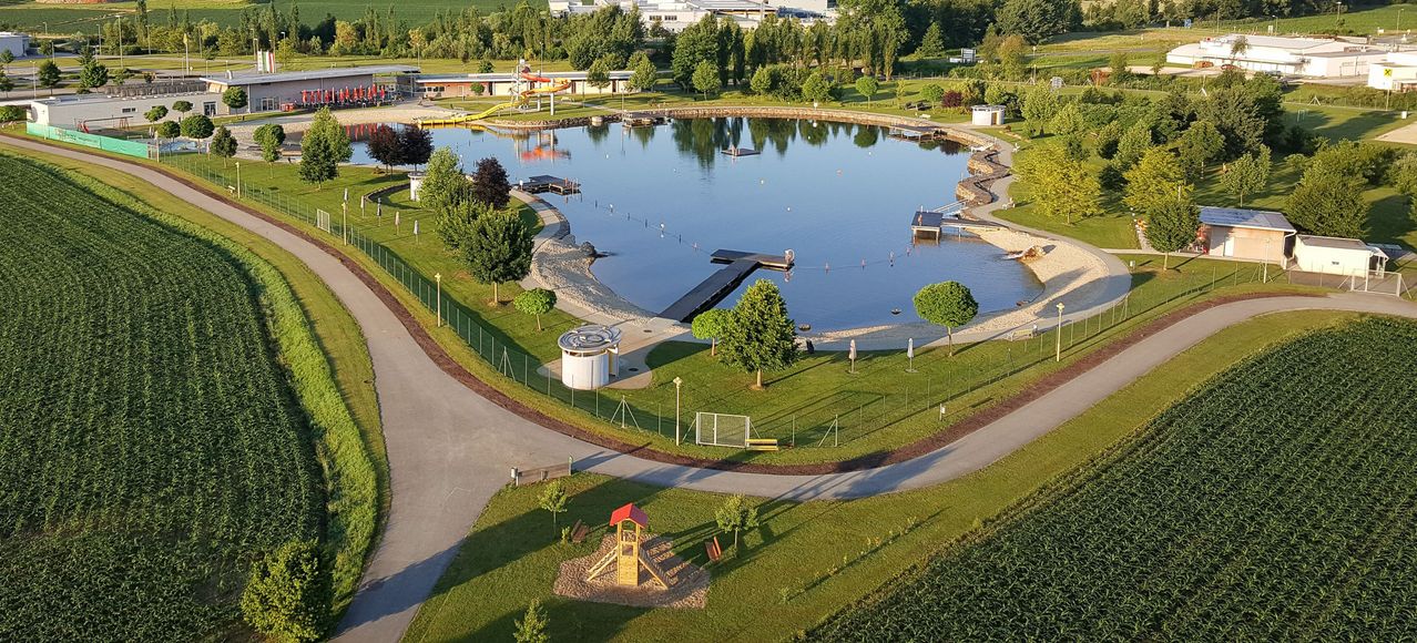 Luftaufnahme eines großen runden Schwimmbeckens mit Rutsche und Anlegestellen, umgeben von üppiger Grünfläche und einem Spielplatz. Gebäude und Bäume in der Ferne.