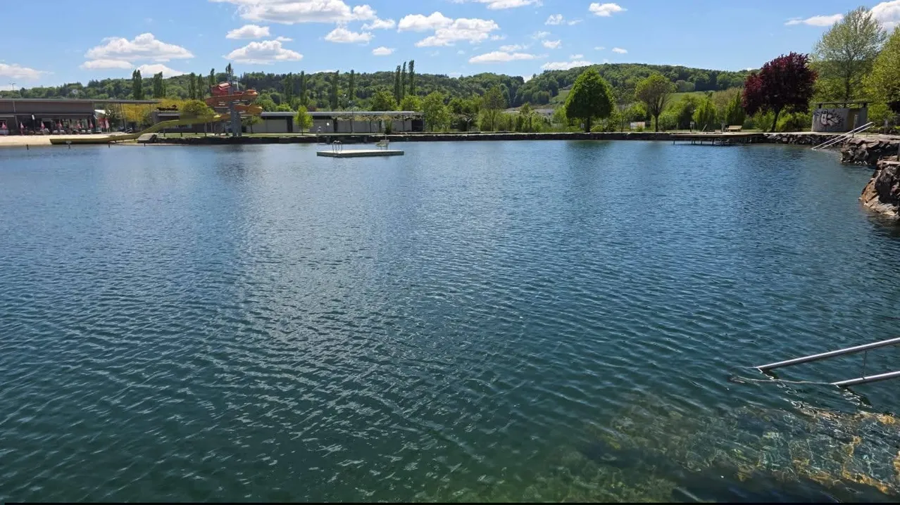Ein ruhiger See mit einem kleinen Steg in der Mitte, umgeben von üppigem Grün und einem entfernten Wasserrutsche unter einem bewölkten Himmel.