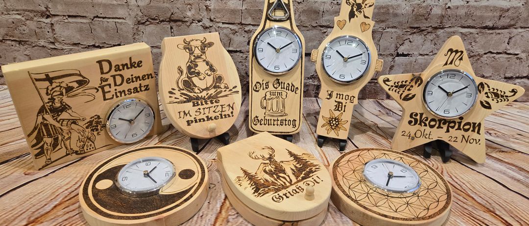 Sieben hölzerne Uhren mit verschiedenen Designs auf einer Holzoberfläche. Eine zeigt eine Kuh und Text auf Deutsch. Eine andere hat ein Reh und eine Blume. Eine dritte hat einen Flaschenöffner und Text. Die vierte zeigt einen Stern und Text. Die fünfte hat eine Blume und Text. Die sechste hat ein Herz und Text. Die siebte zeigt ein Yin-Yang-Symbol und Text.