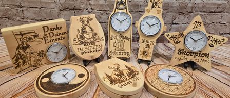 Sieben hölzerne Uhren mit verschiedenen Designs auf einer Holzoberfläche. Eine zeigt eine Kuh und Text auf Deutsch. Eine andere hat ein Reh und eine Blume. Eine dritte hat einen Flaschenöffner und Text. Die vierte zeigt einen Stern und Text. Die fünfte hat eine Blume und Text. Die sechste hat ein Herz und Text. Die siebte zeigt ein Yin-Yang-Symbol und Text.