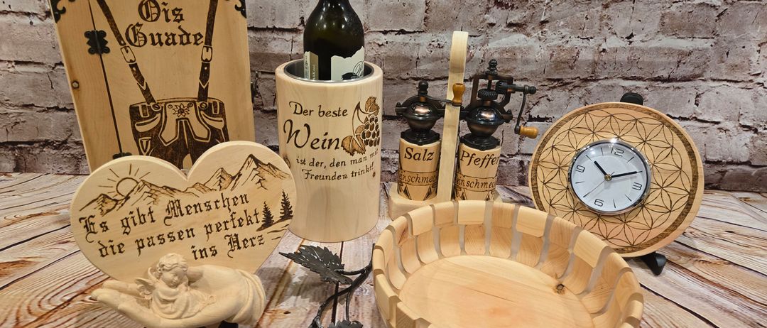 Ein hölzerner Flaschenkühler mit einer Flasche Wein, eine hölzerne Pfeffermühle und eine herzförmige Plakette mit einem Engel darauf stehen auf einem Holztisch.