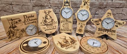 Verschiedene Holzuhren mit unterschiedlichen Designs auf einer Holzoberfläche ausgestellt. Eine zeigt eine Kuh, eine andere ein Reh, und andere haben einzigartige Phrasen wie Danke für Deinen Einsatz.