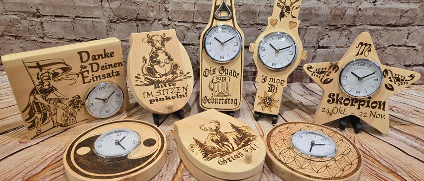 Verschiedene Holzuhren mit unterschiedlichen Designs auf einer Holzoberfläche ausgestellt. Eine zeigt eine Kuh, eine andere ein Reh, und andere haben einzigartige Phrasen wie Danke für Deinen Einsatz.